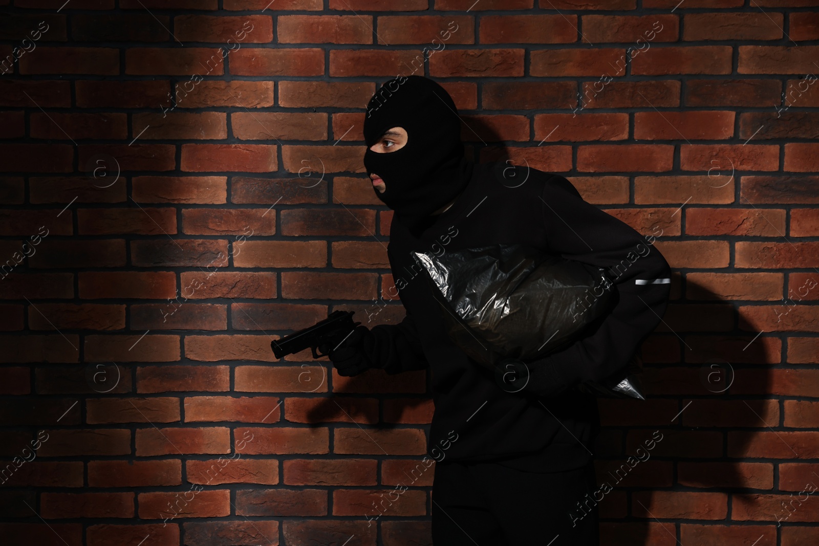 Photo of Thief in balaclava with gun and bag against red brick wall. Space for text