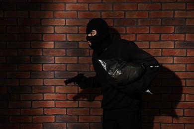 Photo of Thief in balaclava with gun and bag against red brick wall. Space for text