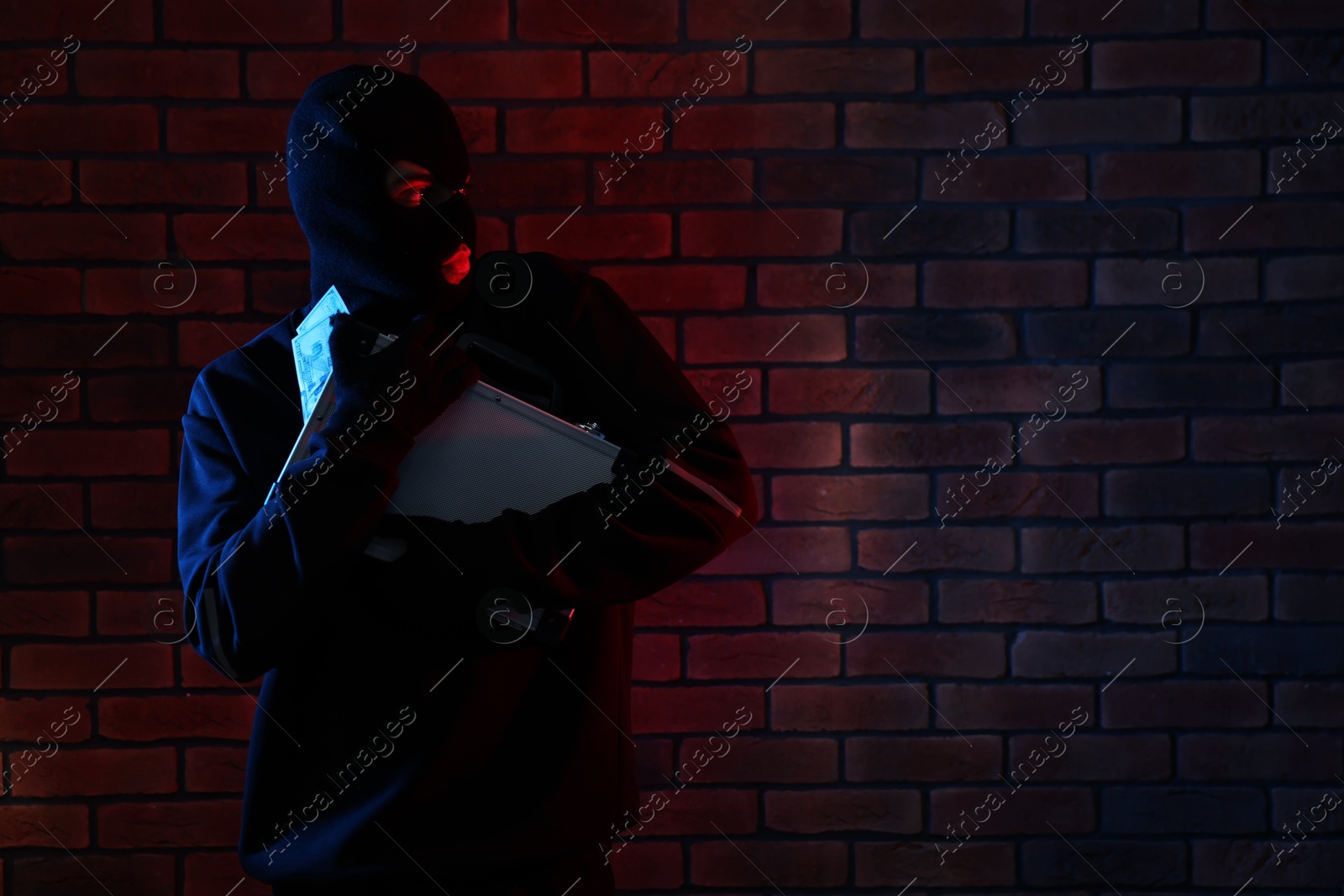 Photo of Thief in balaclava with briefcase of money in color lights against red brick wall. Space for text