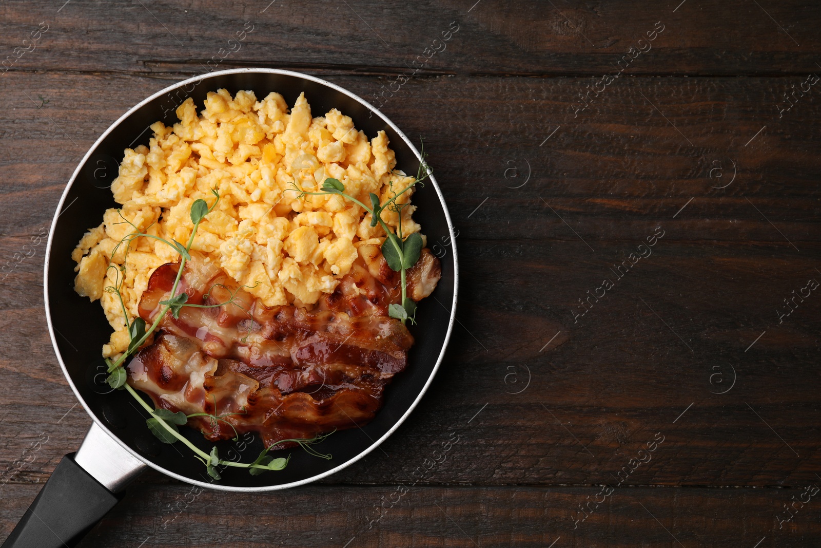 Photo of Delicious scrambled eggs with bacon in frying pan on wooden table, top view. Space for text