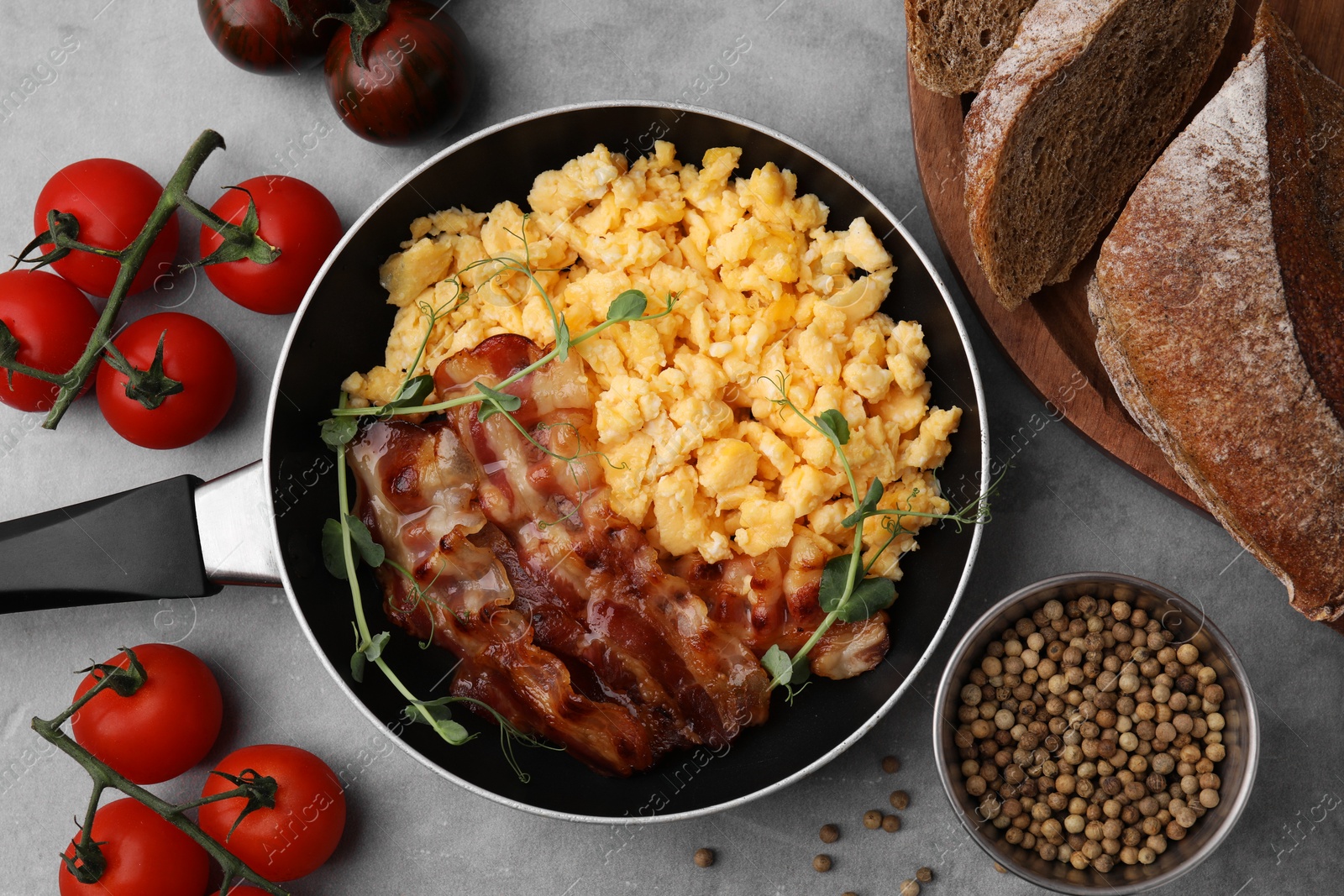 Photo of Delicious scrambled eggs with bacon in frying pan and ingredients on grey table, flat lay