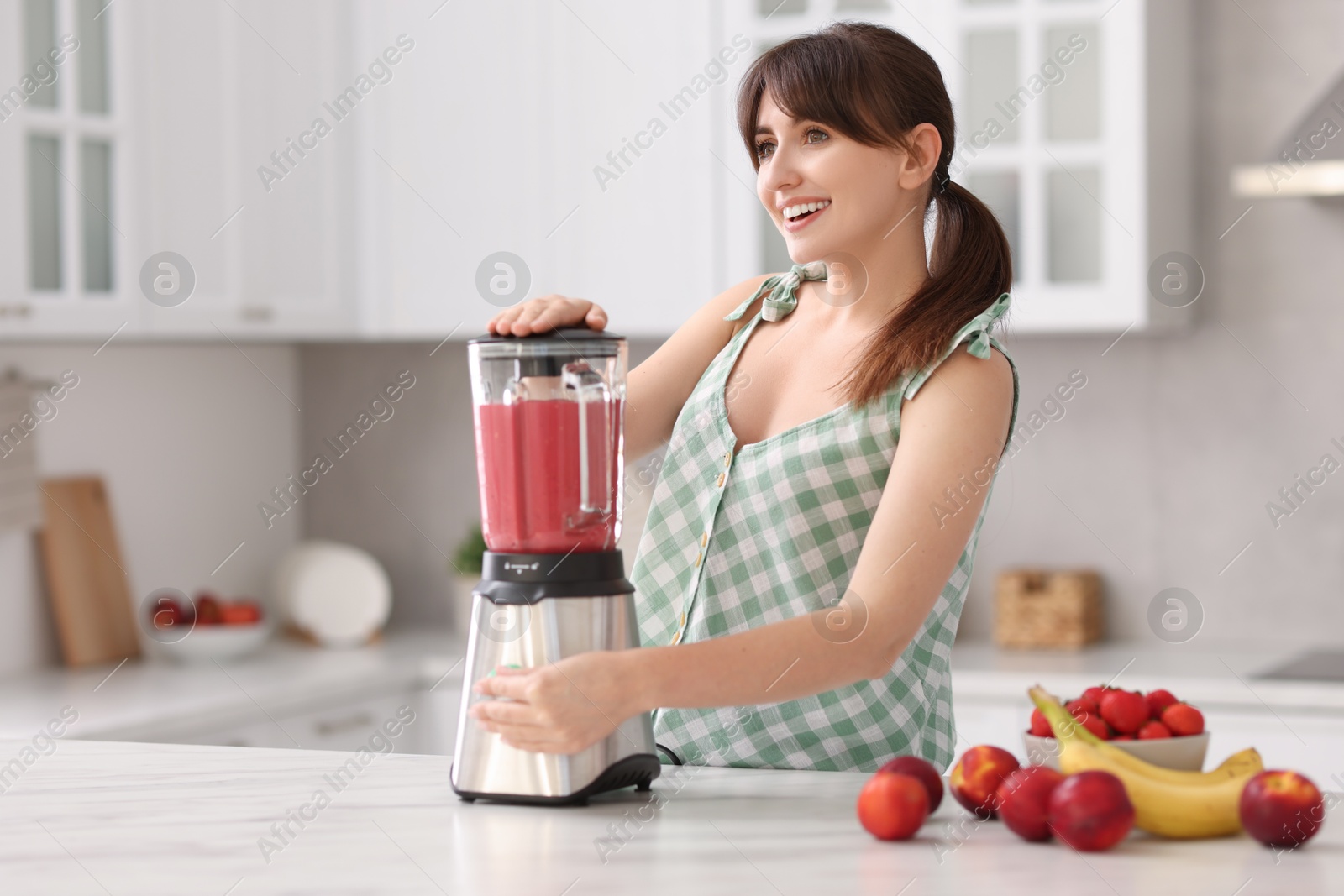 Photo of Young woman making delicious smoothie with blender at white marble table in kitchen