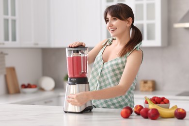 Young woman making delicious smoothie with blender at white marble table in kitchen