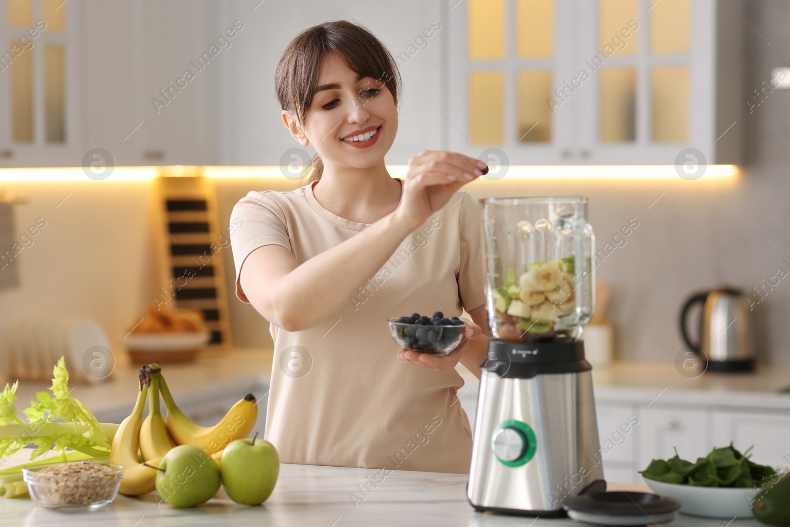 Photo of Young woman making delicious smoothie with blender at white marble table in kitchen