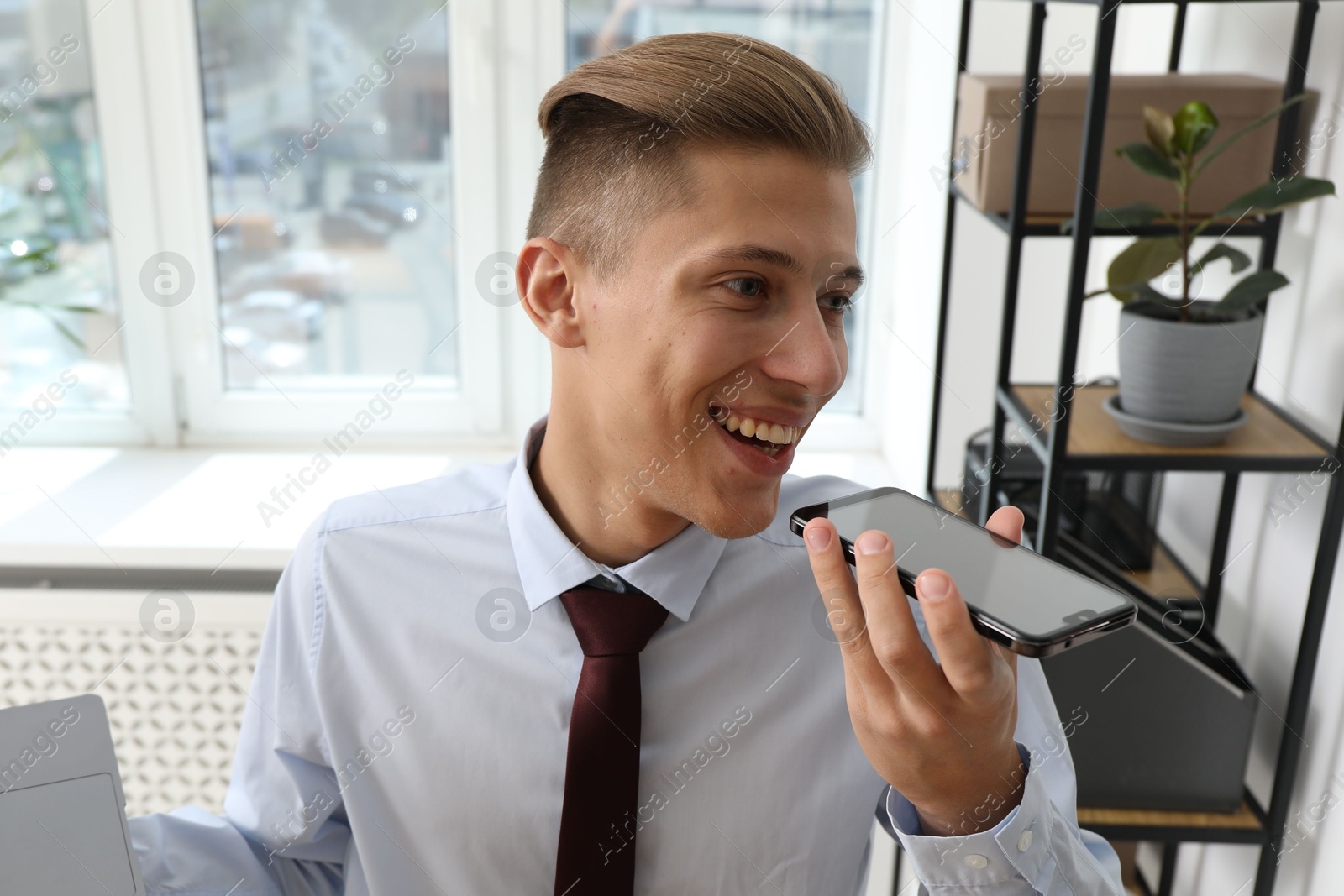 Photo of Young man recording voice message via smartphone in office