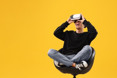 Photo of Happy young man with virtual reality headset sitting on chair against yellow background, space for text