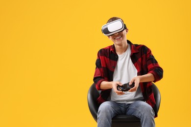 Photo of Happy young man with virtual reality headset and controller sitting on chair against yellow background, space for text