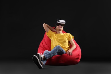Photo of Happy young man with virtual reality headset sitting on bean bag chair against black background