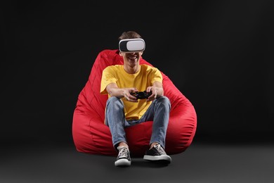 Photo of Happy young man with virtual reality headset and controller sitting on bean bag chair against black background
