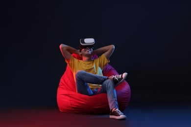 Photo of Happy young man with virtual reality headset sitting on bean bag chair in neon lights against black background