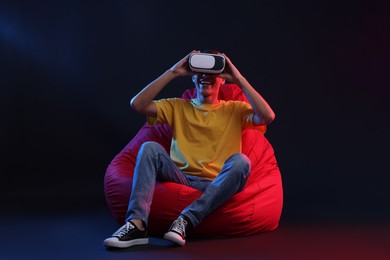 Photo of Happy young man with virtual reality headset sitting on bean bag chair in neon lights against black background