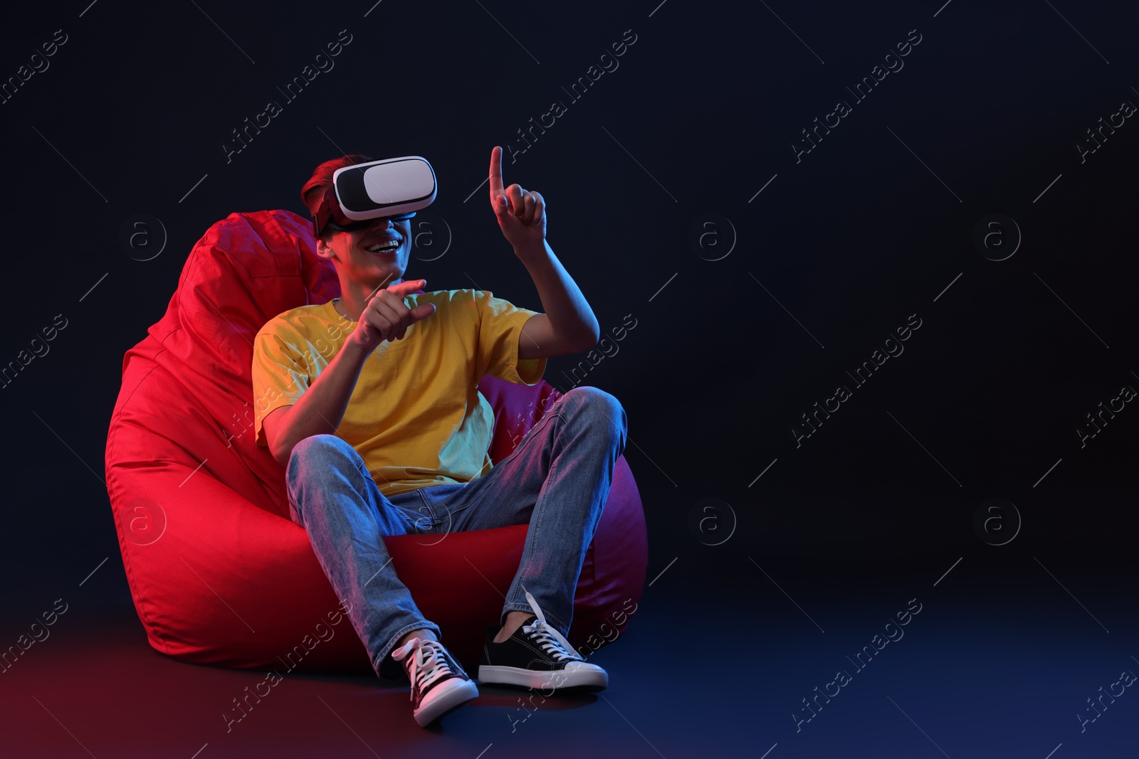 Photo of Happy young man with virtual reality headset sitting on bean bag chair in neon lights against black background, space for text