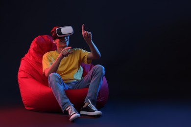 Photo of Happy young man with virtual reality headset sitting on bean bag chair in neon lights against black background, space for text