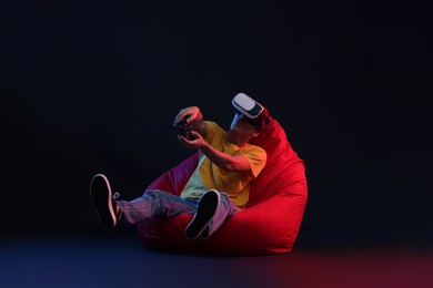 Photo of Emotional young man with virtual reality headset and controller sitting on bean bag chair in neon lights against black background
