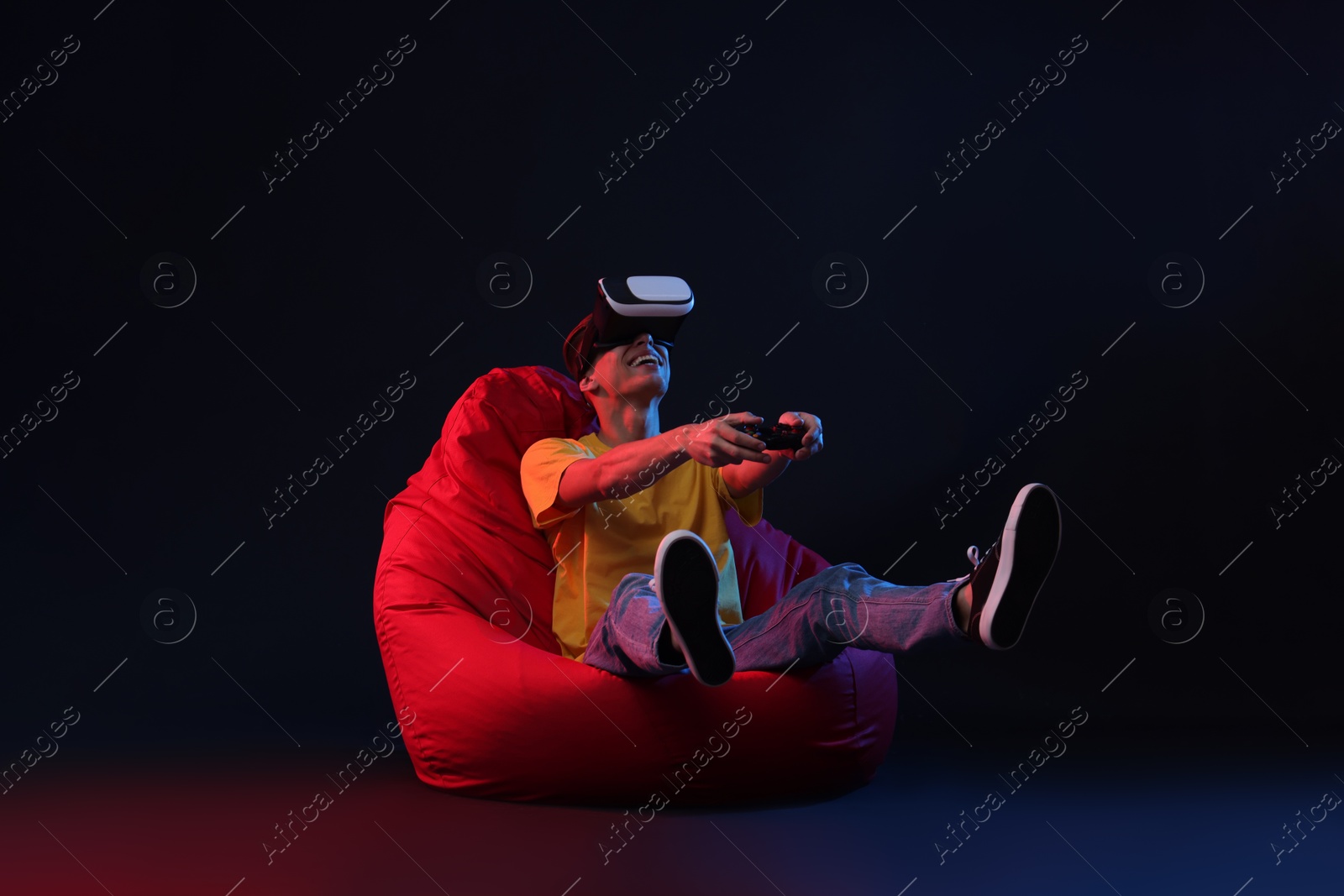 Photo of Happy young man with virtual reality headset and controller sitting on bean bag chair in neon lights against black background