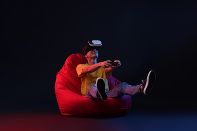 Photo of Happy young man with virtual reality headset and controller sitting on bean bag chair in neon lights against black background