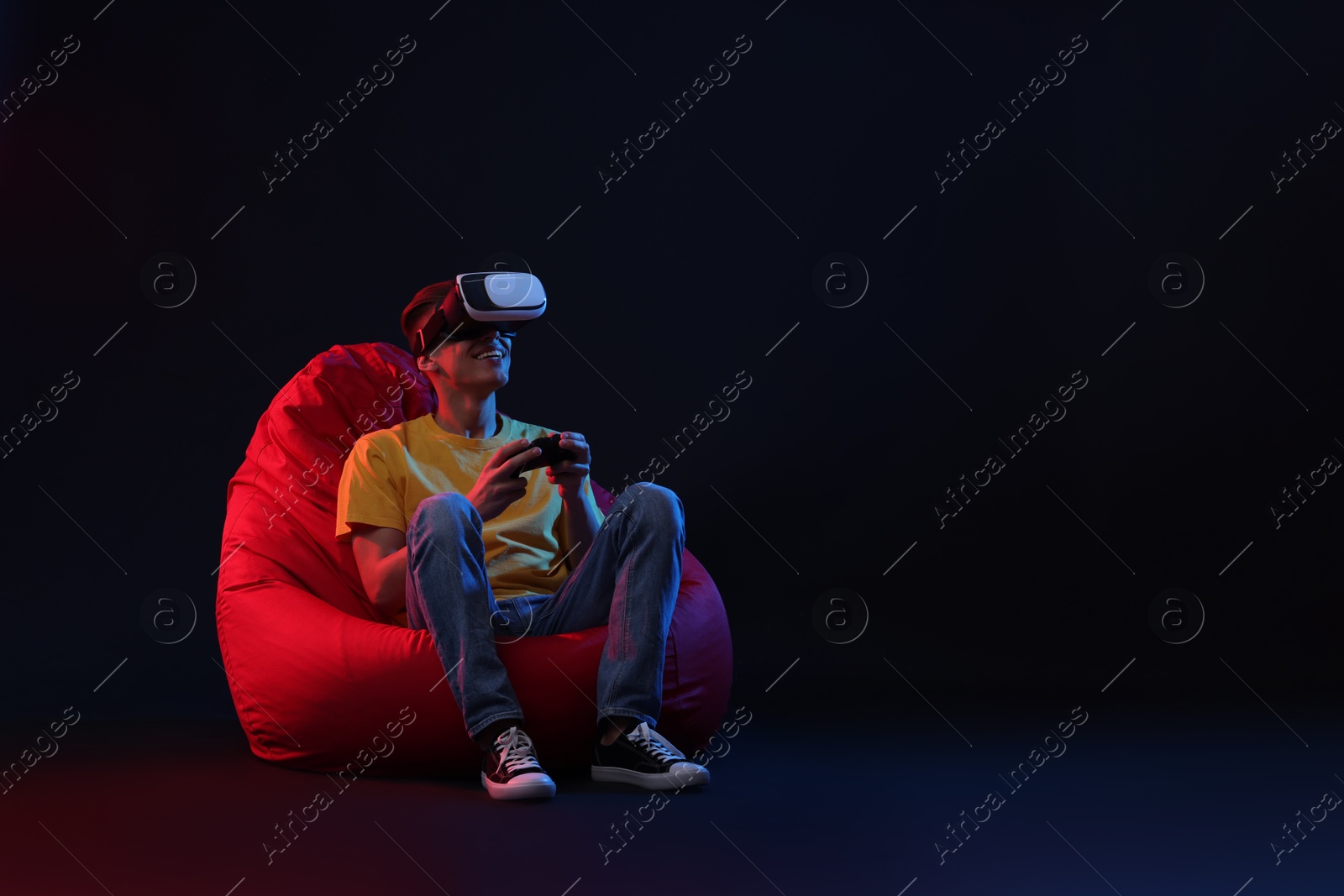 Photo of Happy young man with virtual reality headset and controller sitting on bean bag chair in neon lights against black background, space for text