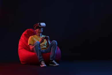 Photo of Happy young man with virtual reality headset and controller sitting on bean bag chair in neon lights against black background, space for text