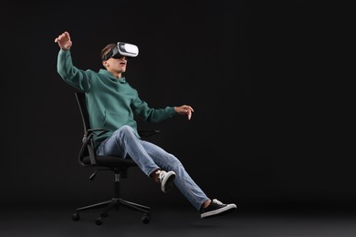 Photo of Emotional young man with virtual reality headset sitting on chair against black background, space for text