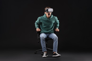 Photo of Emotional young man with virtual reality headset sitting on chair against black background