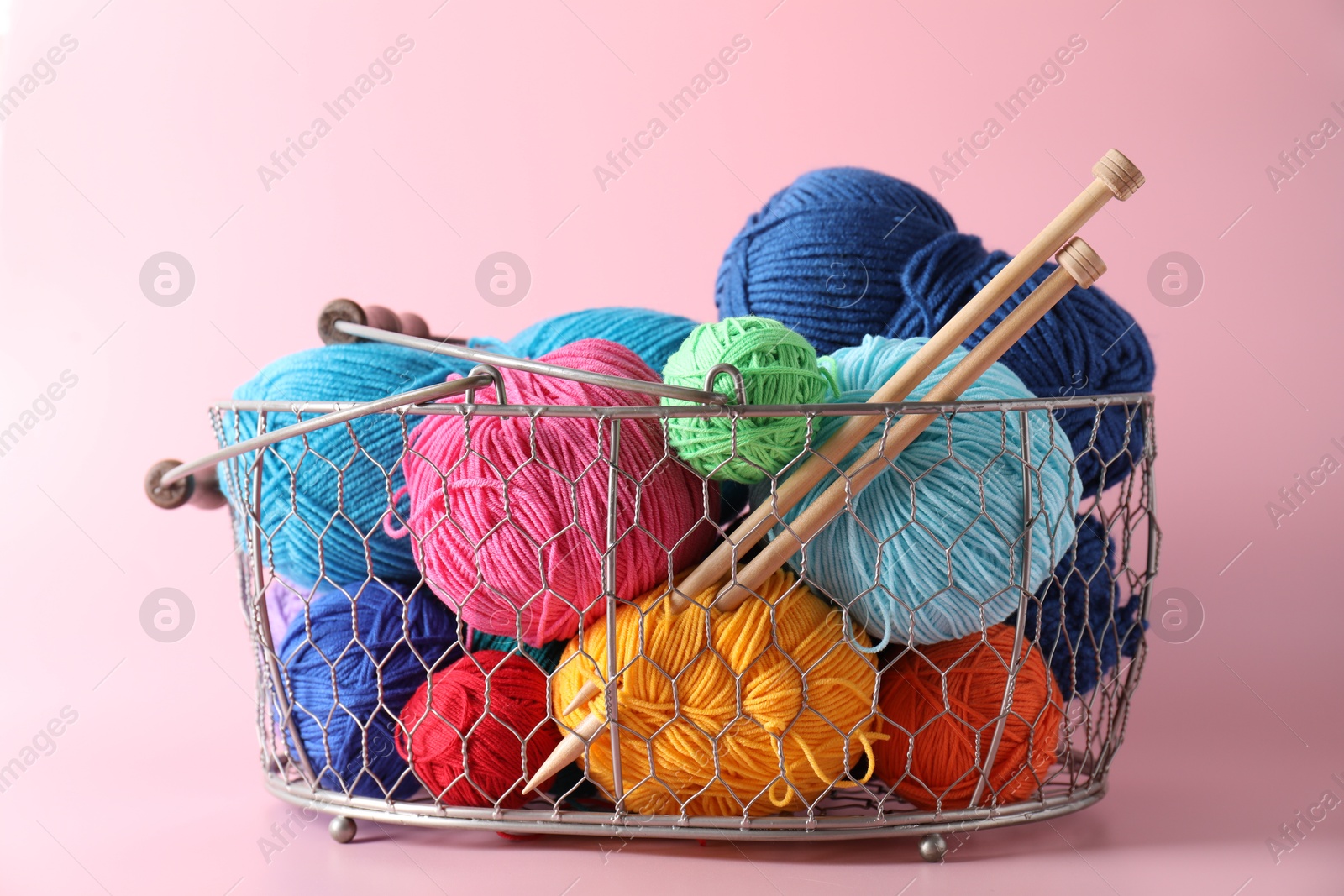 Photo of Metal basket with bright yarns and knitting needles on pink background, closeup