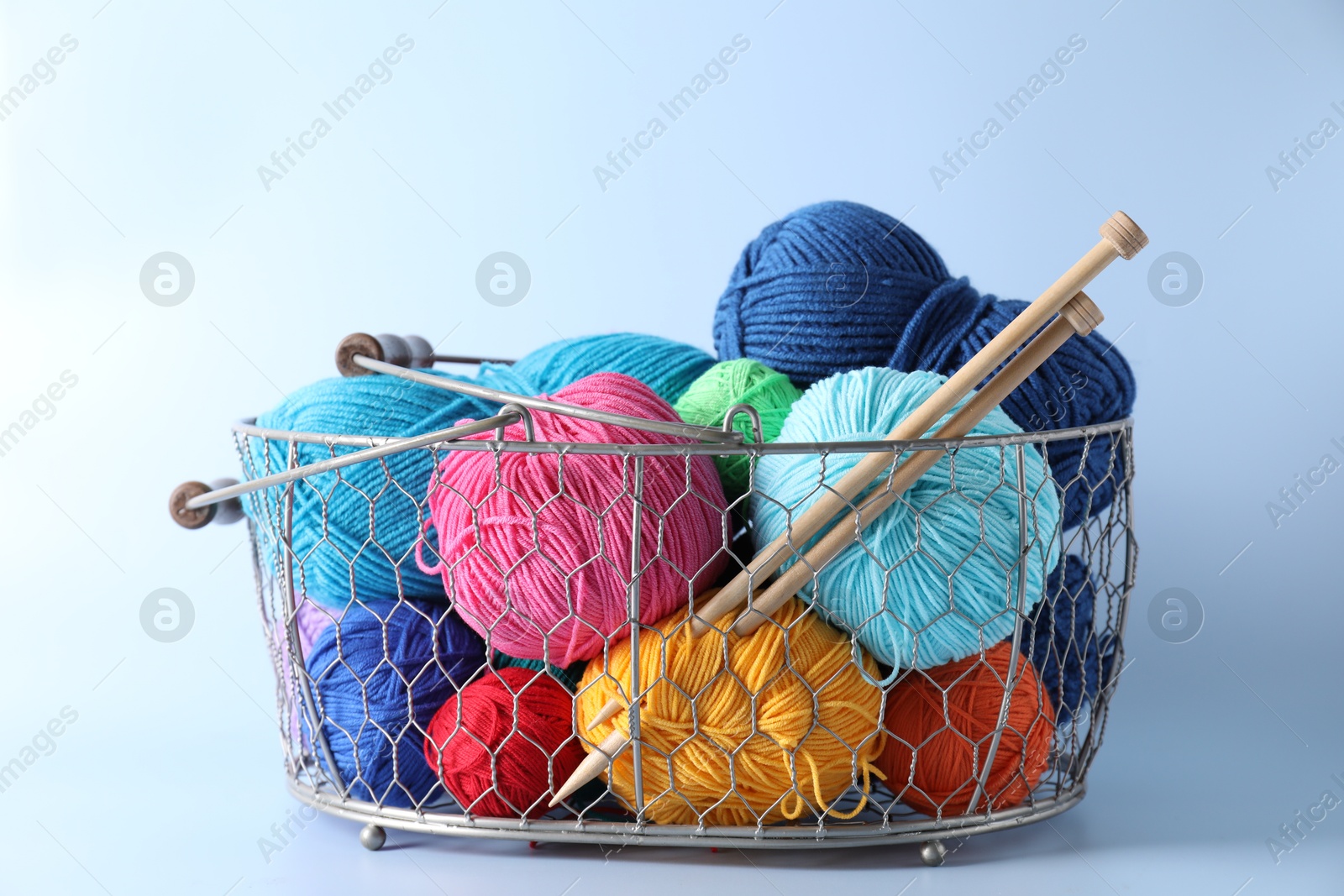 Photo of Metal basket with bright yarns and knitting needles on light blue background, closeup