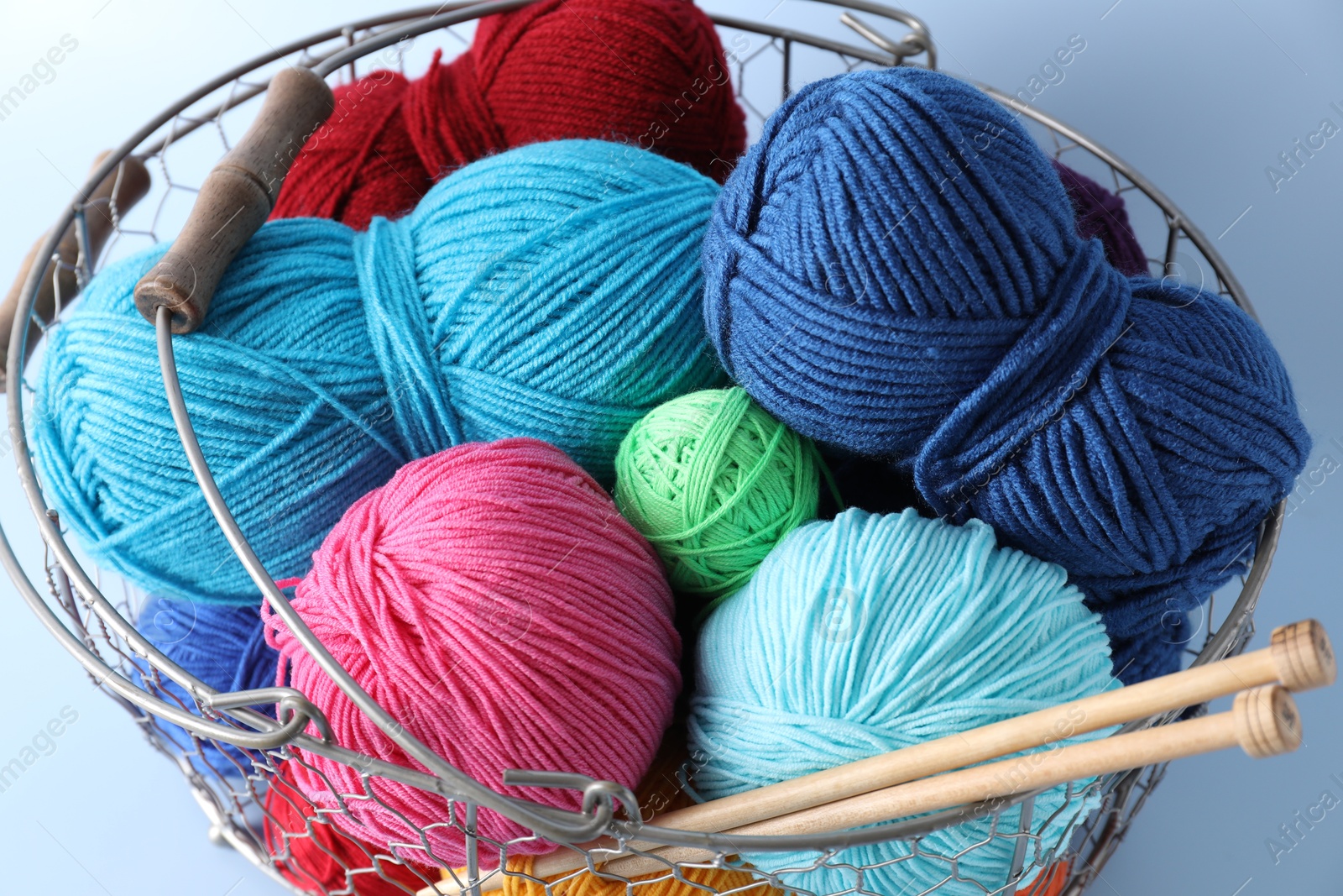 Photo of Metal basket with bright yarns and knitting needles on light blue background, closeup