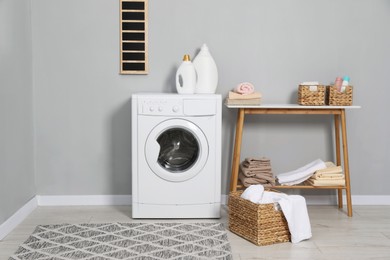 Photo of Washing machine, detergents, basket with laundry and storage bench indoors