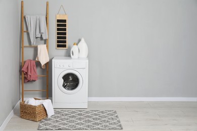 Washing machine, detergents, ladder, basket and laundry near grey wall indoors, space for text