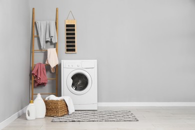 Photo of Washing machine, detergents, ladder, basket and laundry near grey wall indoors, space for text