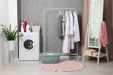 Photo of Washing machine, detergents, baskets, clothing rack and mirror indoors