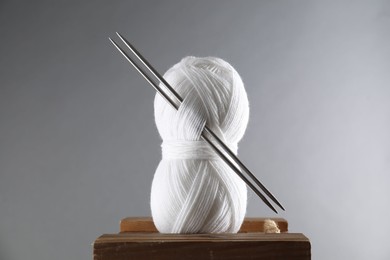 Skein of white yarn and knitting needles on wooden stand against grey background