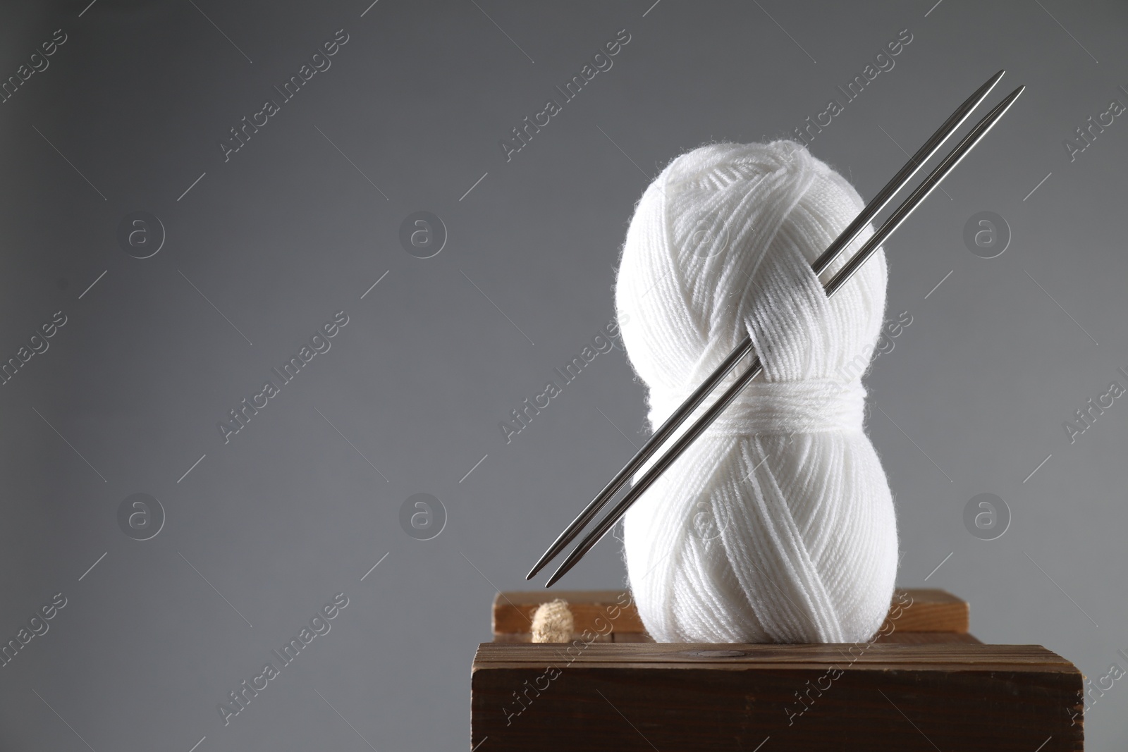 Photo of Skein of white yarn and knitting needles on wooden stand against grey background, space for text