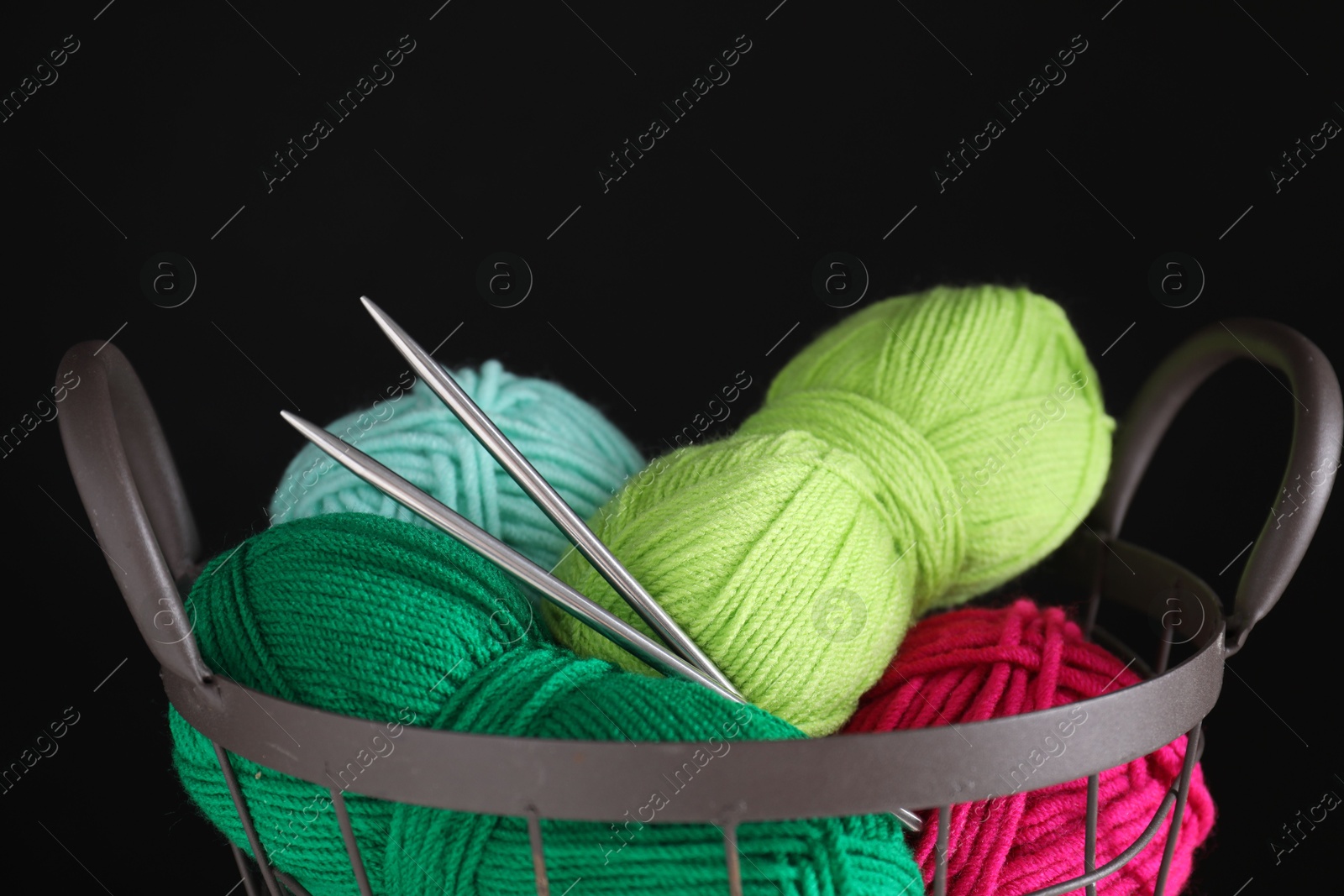 Photo of Many soft skeins of yarn and knitting needles in metal basket on black background, closeup