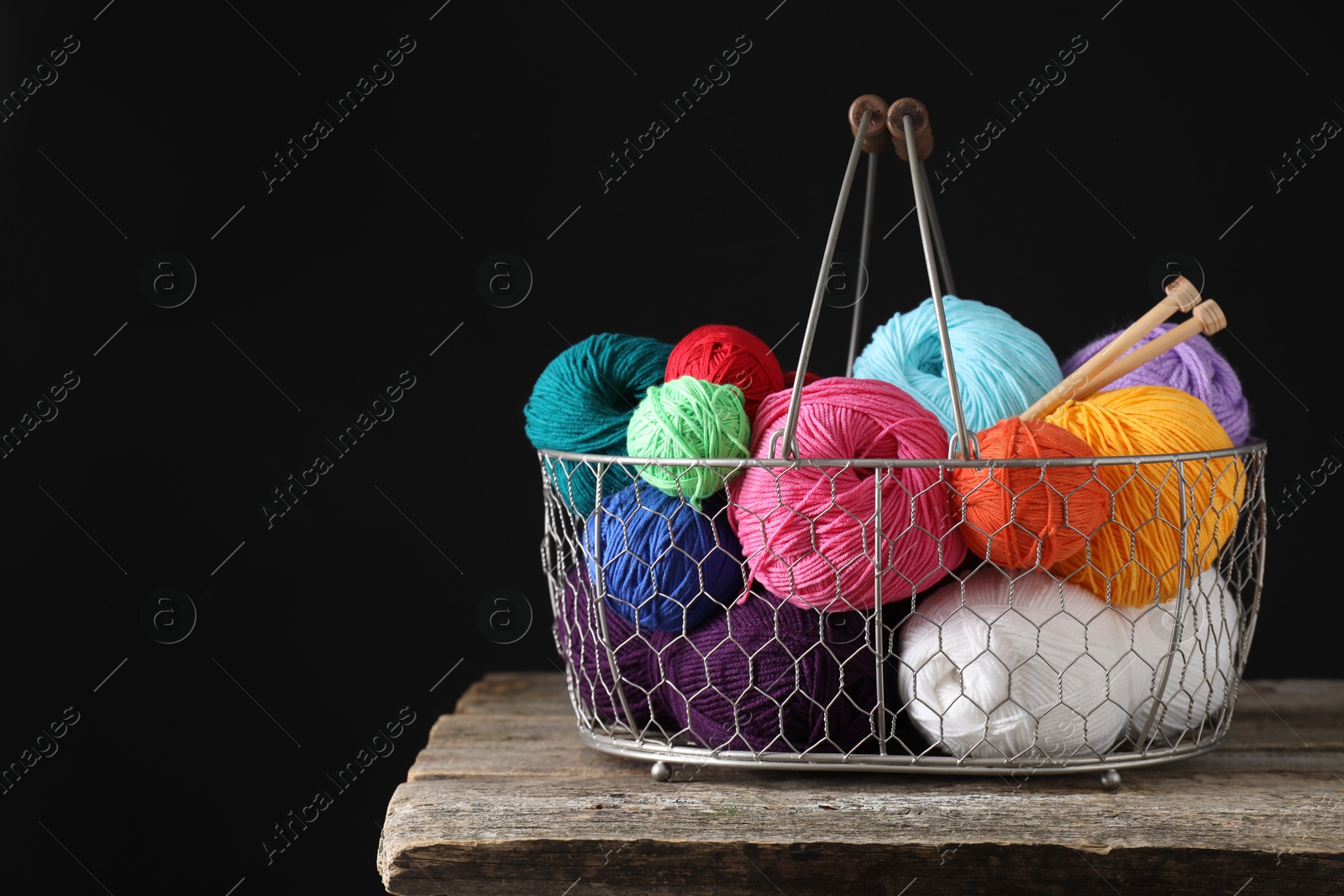 Photo of Many soft skeins of yarn and knitting needles in metal basket on wooden table, space for text