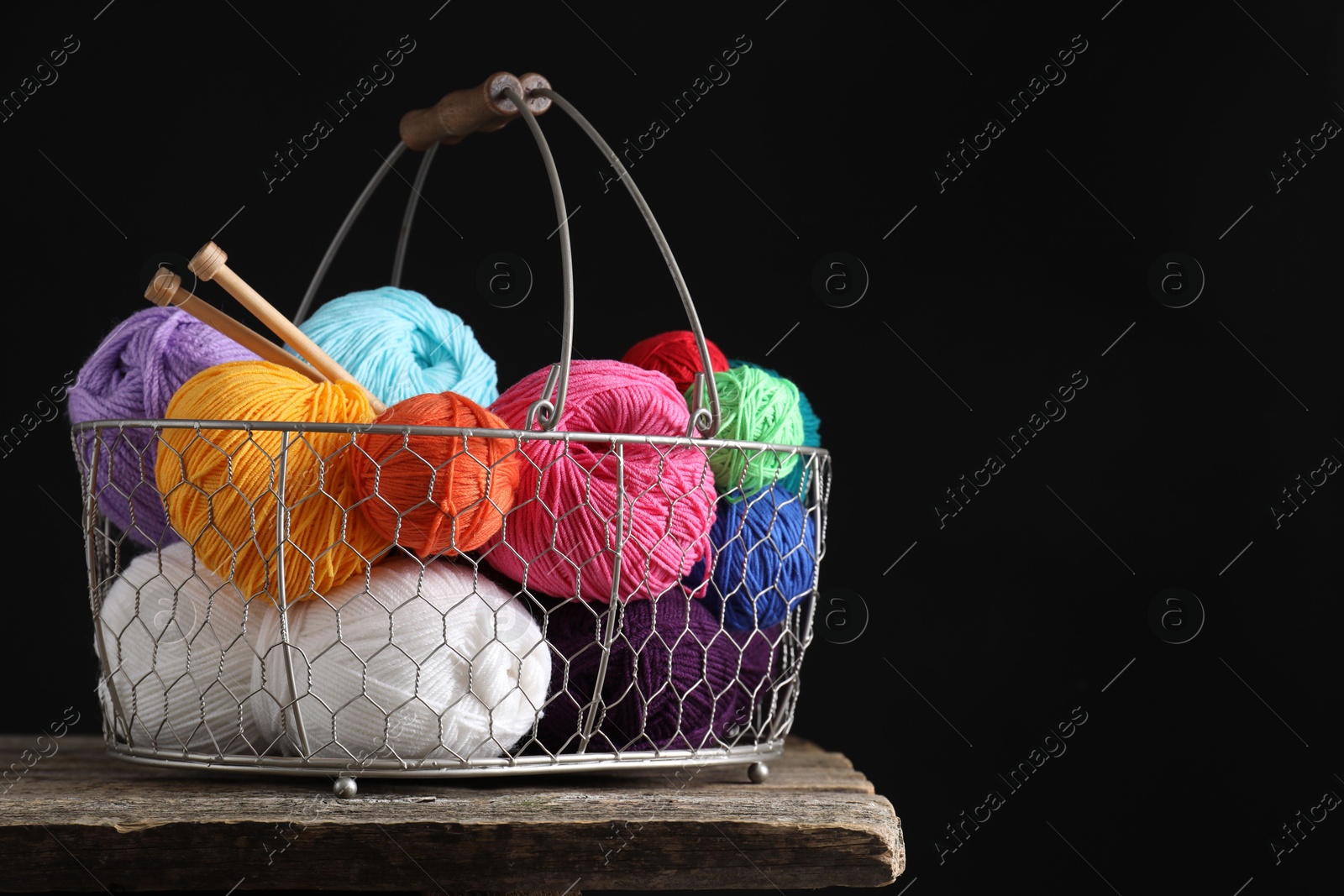 Photo of Many soft skeins of yarn and knitting needles in metal basket on wooden table, space for text