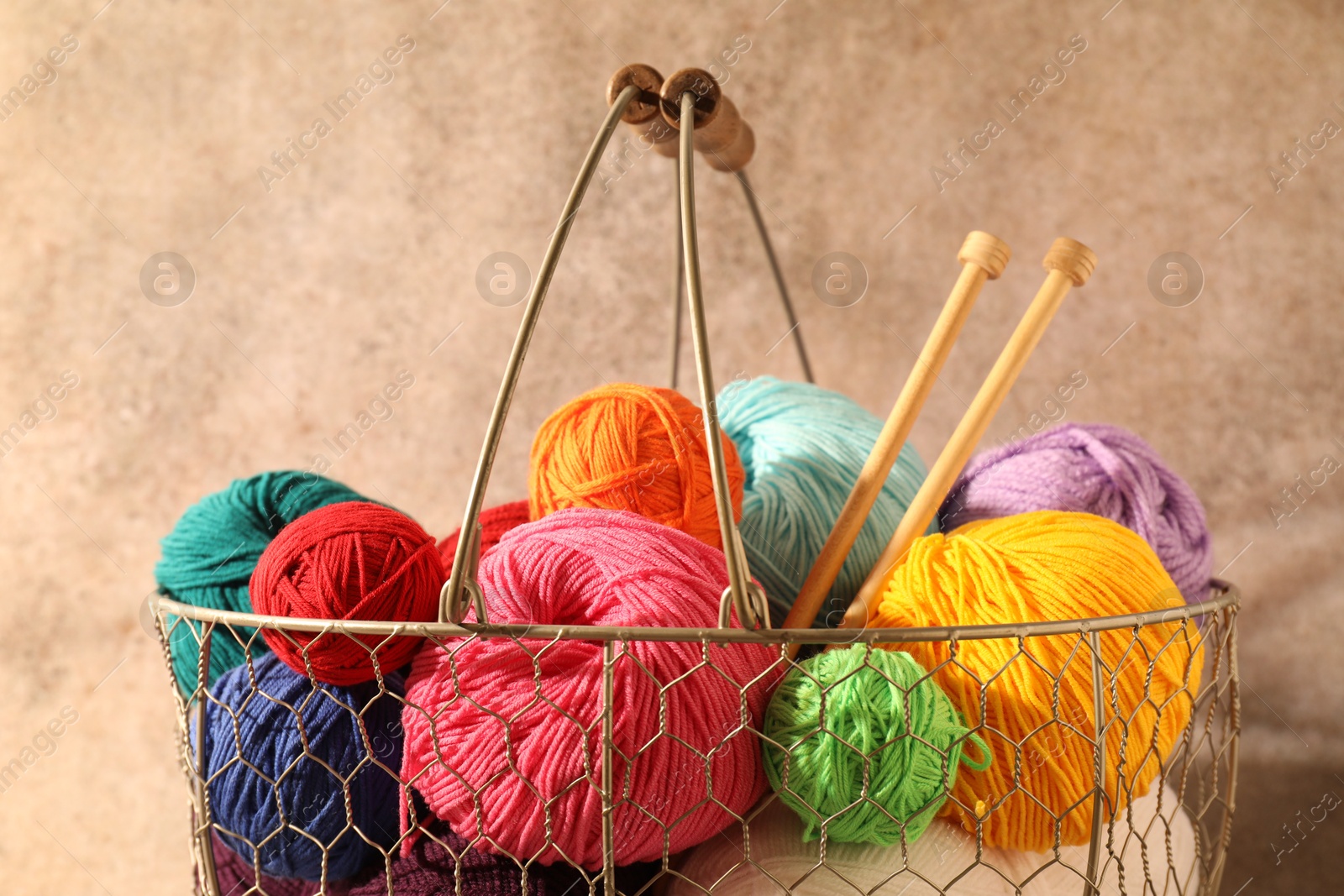 Photo of Many soft skeins of yarn and knitting needles in metal basket on light brown background, closeup