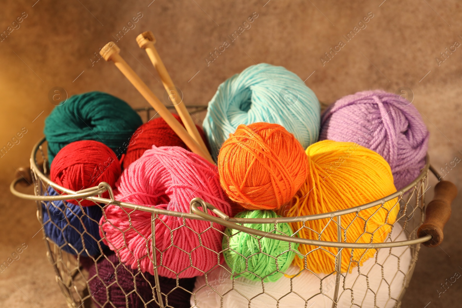 Photo of Many soft skeins of yarn and knitting needles in metal basket on light brown background, closeup