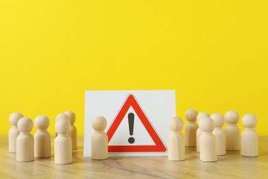 Wooden human figures among warning road sign on table against yellow background