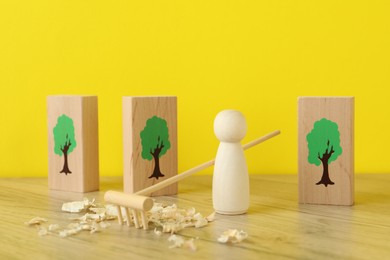 Photo of Wooden human figure with rake and sawdust among planks with drawn trees on table against yellow background