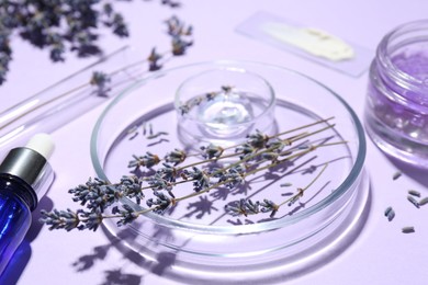 Photo of Petri dishes with lavender flowers and cosmetic products on violet background, closeup