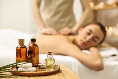 Photo of Aromatherapy. Woman receiving back massage in spa salon, focus on bottles of essential oils, burning candle and stones