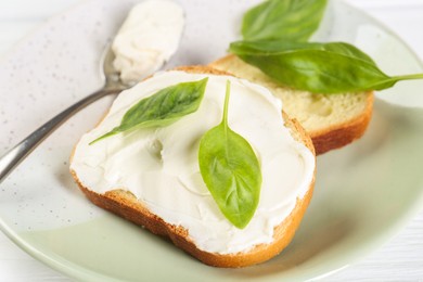 Pieces of bread with cream cheese and basil leaves on white wooden table, closeup