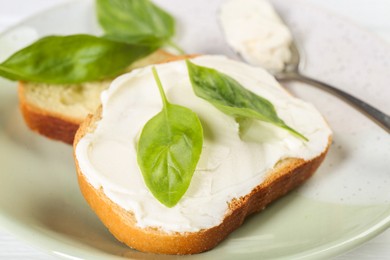 Pieces of bread with cream cheese and basil leaves on white wooden table, closeup