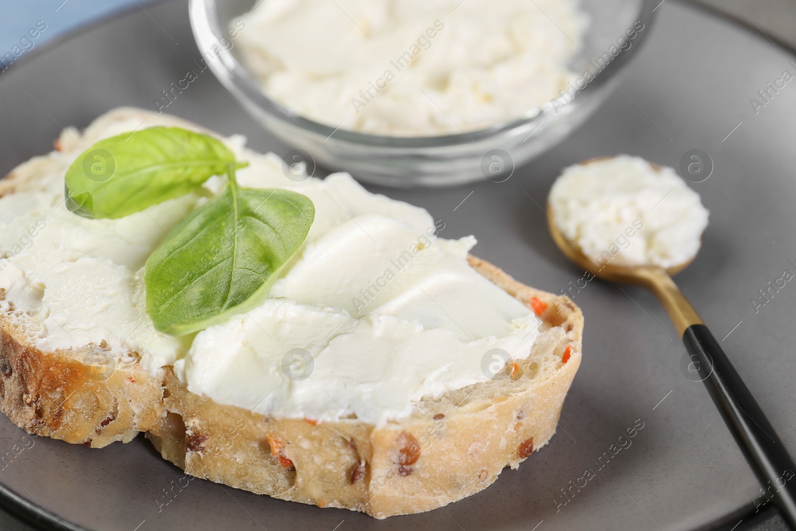 Photo of Piece of bread with cream cheese and basil leaves on gray plate, closeup