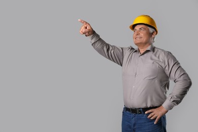 Engineer in hard hat pointing at something on grey background, space for text