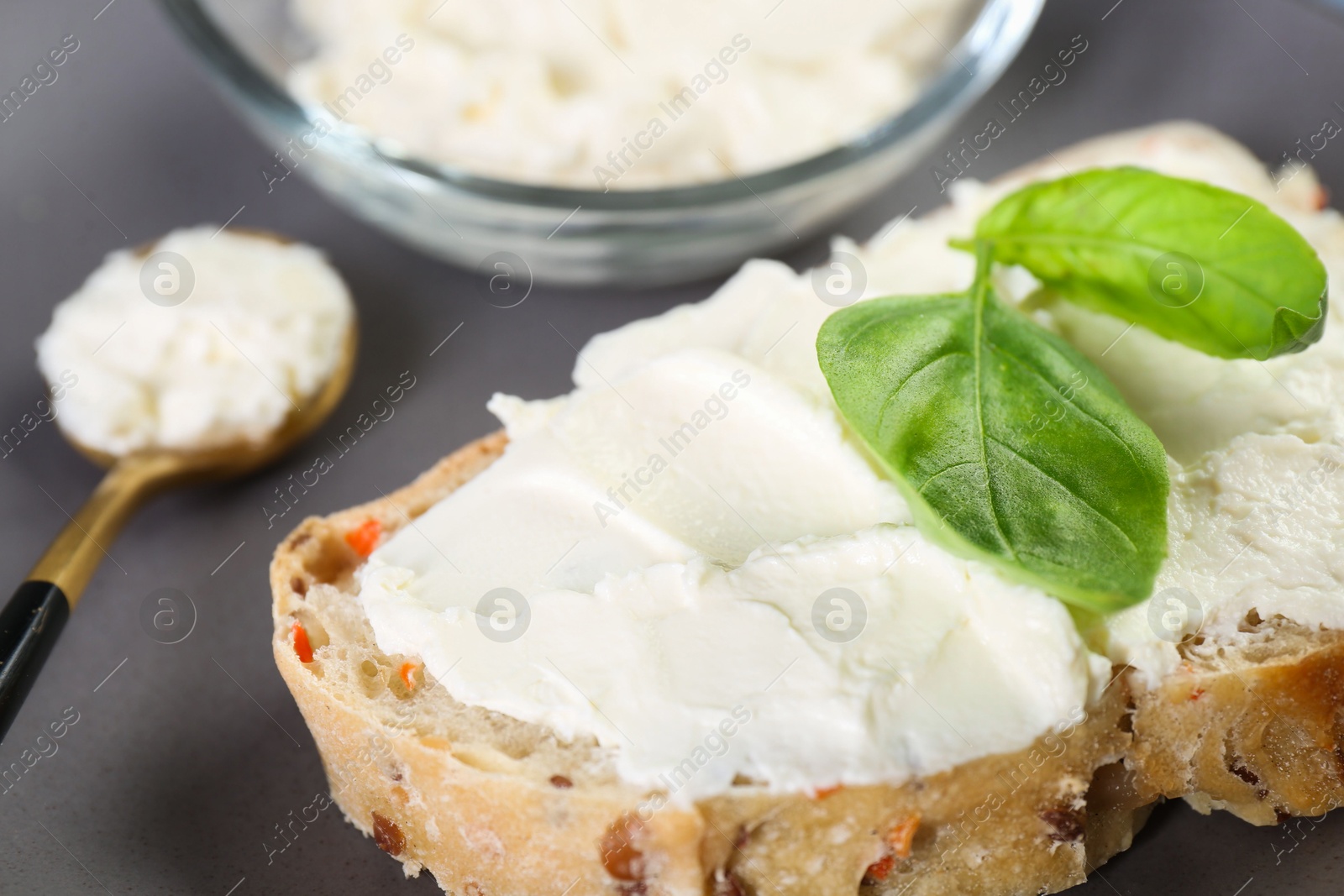 Photo of Piece of bread with cream cheese and basil leaves on gray table, closeup