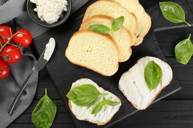 Pieces of bread with cream cheese and basil served on black wooden table, flat lay