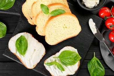 Pieces of bread with cream cheese and basil served on black wooden table, flat lay