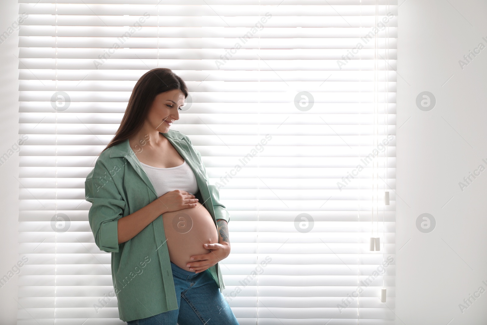 Photo of Beautiful pregnant woman near window blinds at home, space for text